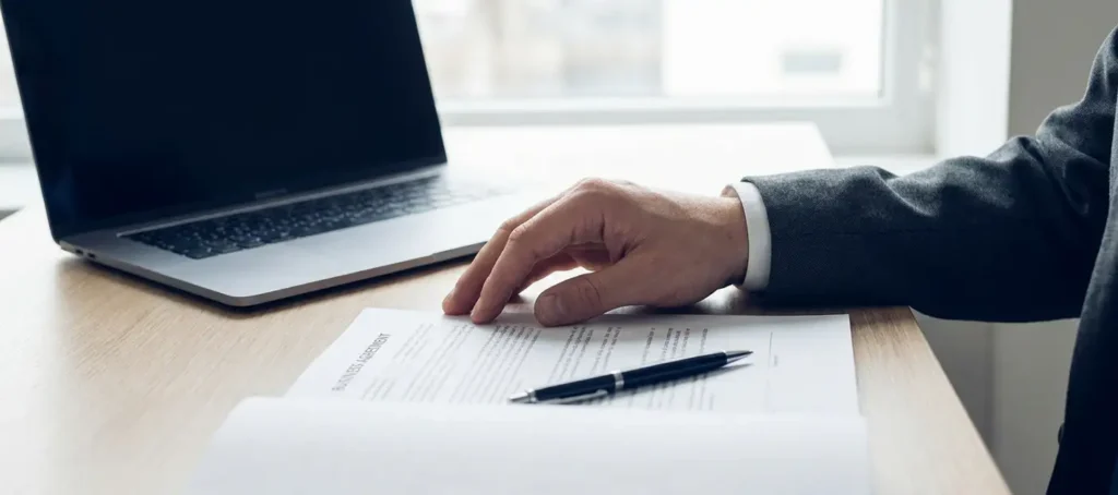 Hand op een gedrukt contract naast een laptop op een modern bureau, met een pen over de pagina's.