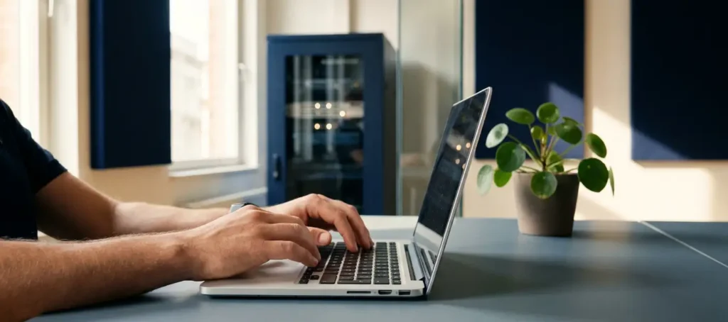 Data-engineer typt op laptop in modern kantoor met serverrack op achtergrond en kantoorplant op bureau.