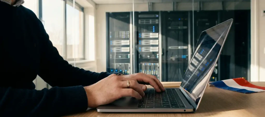 IT-professional typt op laptop met serverruimte achter glazen wand en Nederlandse vlag op bureau.