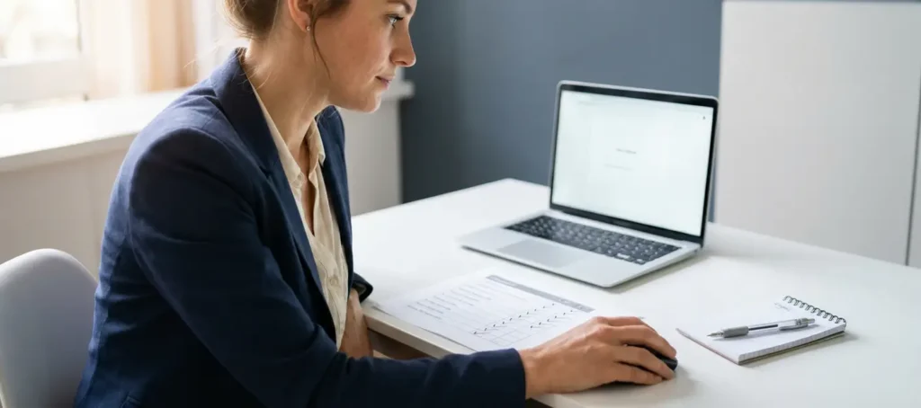 A customer service representative carefully examines a laptop and a checklist on a white desk while supervising an automated process.