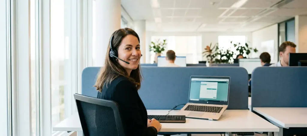 Klantenservicemedewerker met headset aan modern bureau in open kantoor, laptop en softphone zichtbaar, natuurlijk daglicht.