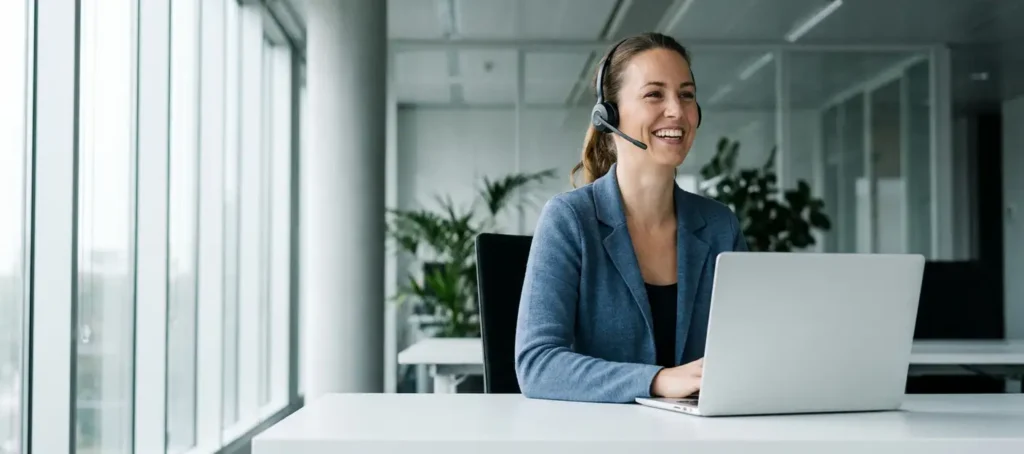 Glimlachende klantenservicemedewerker met headset aan modern bureau in licht kantoor met laptop en grote ramen.