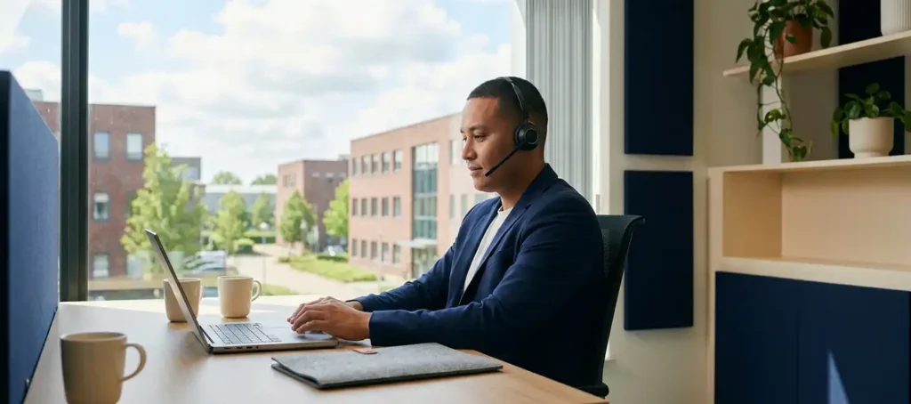 Klantenservicemedewerker met headset achter laptop in modern Nederlands kantoor met groot raam en natuurlijk daglicht.