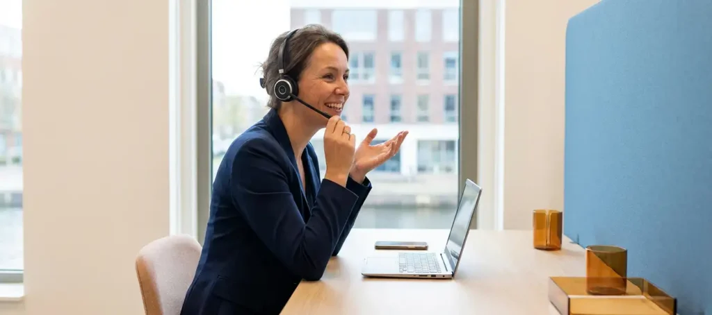 Klantenservicemedewerker met headset aan modern bureau in Nederlands kantoor, laptop en smartphone zichtbaar, natuurlijk licht.