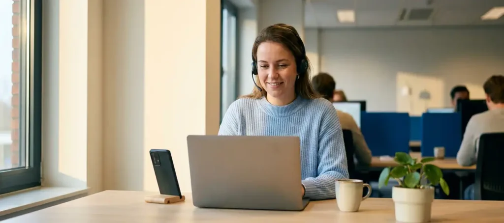 Klantenservicemedewerker met headset glimlacht aan modern bureau met laptop en smartphone in licht Nederlands kantoor.