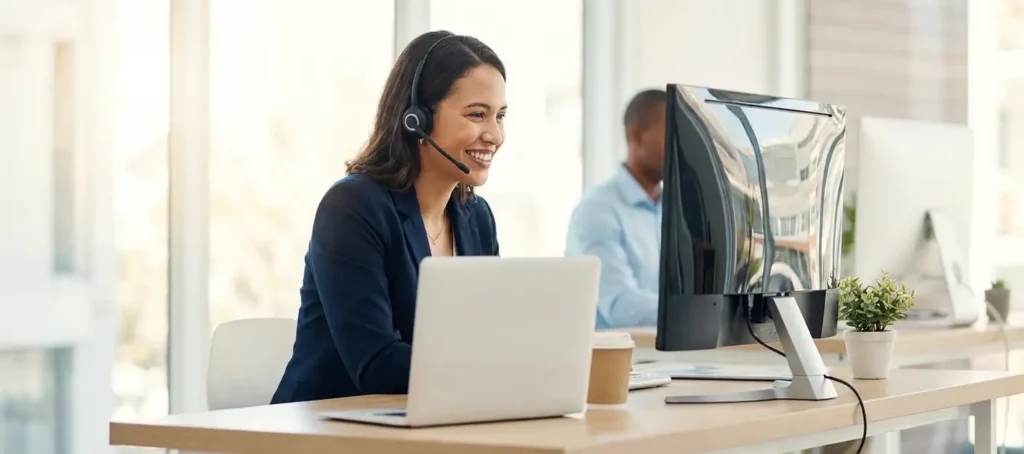 A customer service representative wearing a headset smiles at a monitor in a modern office, with a coworker in the background.