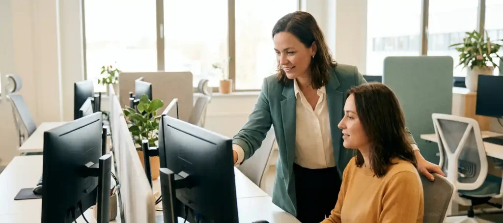 Teamleider begeleidt collega bij werkplek in modern contactcenter met natuurlijk licht en open kantooromgeving.