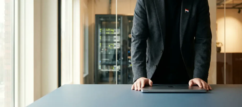 Professional leunend over modern bureau met gesloten laptop, serverrack zichtbaar achter glazen wand, Nederlandse vlagspeld op revers.