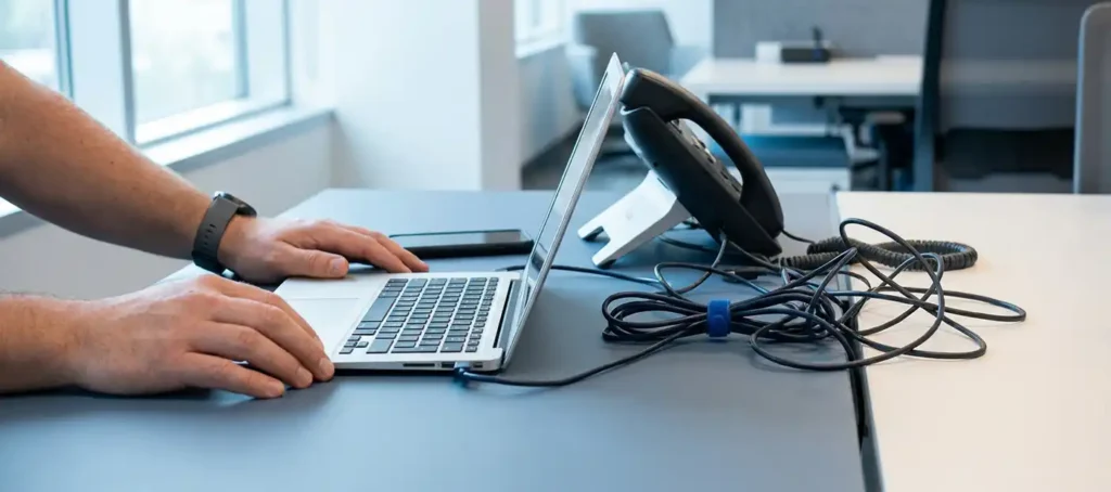 Ontwikkelaarshanden bij laptop op modern bureau met desk phone, smartphone en kabels die communicatie-integratie symboliseren.