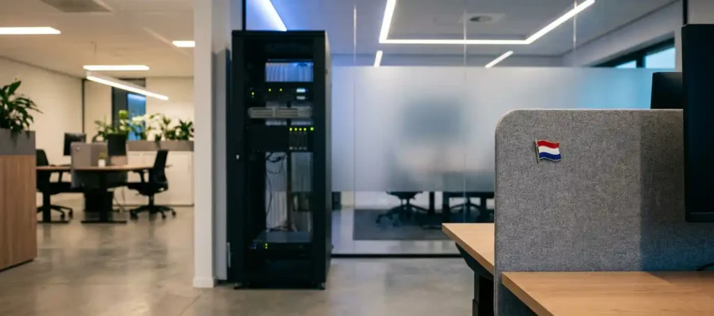 Serverrack in modern Nederlands kantoor met glazen wand, Nederlandse vlaggenpin op bureau en gepolijste betonvloer.