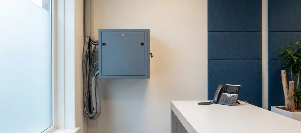 Wandgemonteerd serverrack naast een VoIP-bureautelefoon op wit bureau in modern Nederlands kantoor.