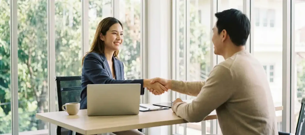 A customer service representative and a satisfied customer shake hands at a modern desk with a laptop and a cup of coffee in a bright office.