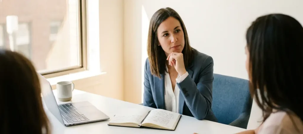 A business professional listens attentively to a client at a modern desk with a notebook and laptop, bathed in warm, natural light.